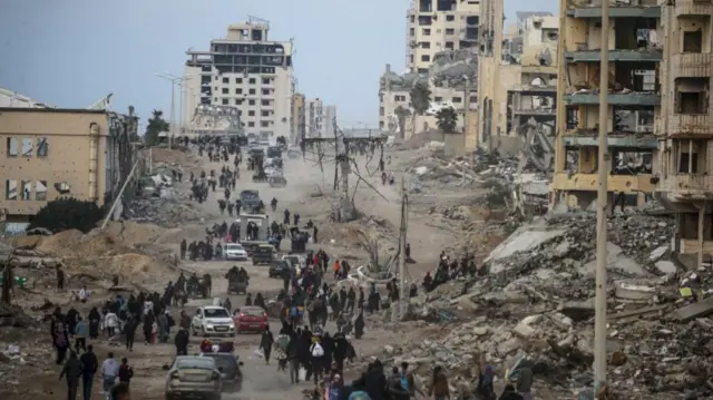 People walking along a road in Gaza City with crumbling buildings and rubble on both sides