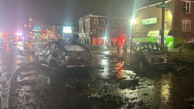 Burnt out cars on road covered with debris, residential buildings and shops in background