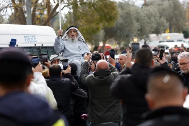 A man in a headscarf and a grey jumper being held above a crowd as many take pictures