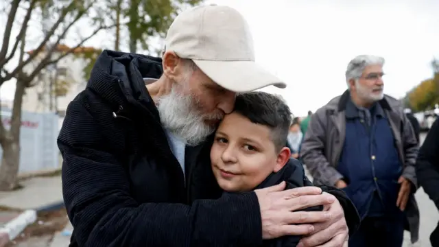 A man with white beard and cap on hugs a teenage boy who is smiling