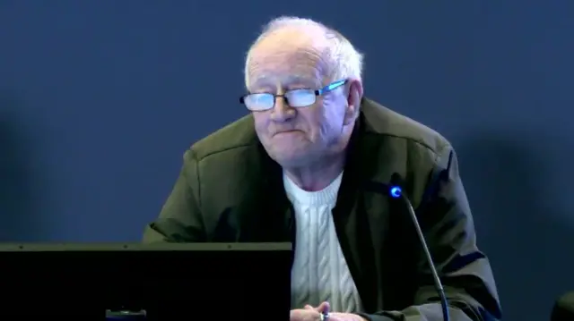 A man sits behind a microphone and a computer screen, he has a green jacket and cream jumper. He is balding with wispy grey hair and glasses. He is sitting in front of a blue background.