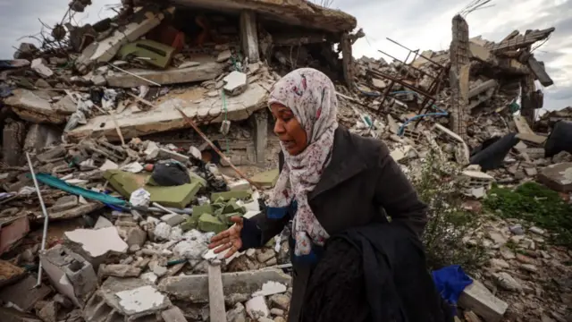 Woman walks across ruins of bombed out home in Gaza. She's wearing a dark coat and floral head scarf