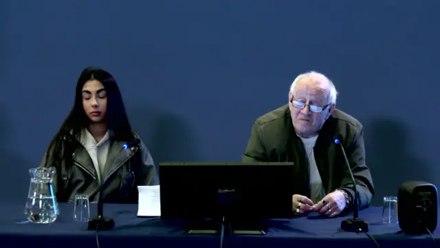 An older man with balding grey hair is sitting behind a computer monitor and a microphone. He is wearing a dark green jacket and a cream jumper. He is sitting in front of a blue background.