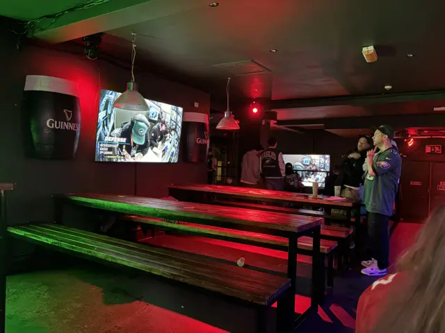 A pub with empty benches and a few American football fans standing in front of a TV