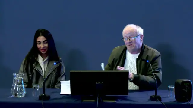 A man with balding grey hair sits to the right behind a computer monitor and microphone; to his left is a young woman with shoulder-length black hair - they are both sitting in front of a blue background