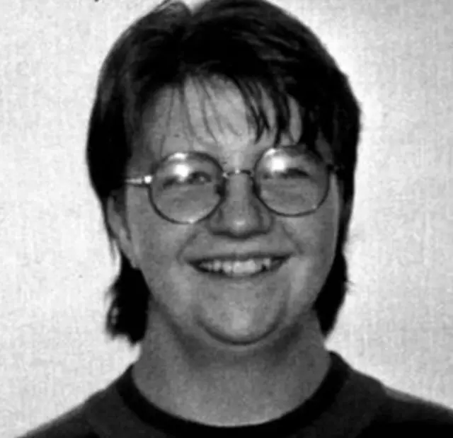 A black and white image of a young woman. She has short hair and round glasses. A plain wall is in the background