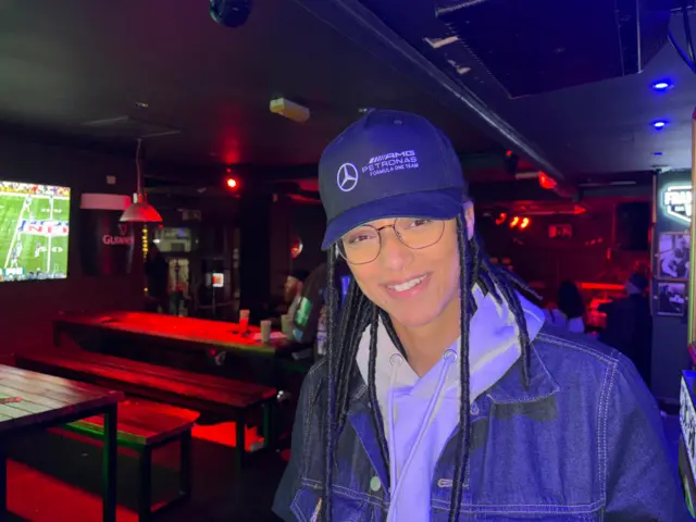 Kenya has dreadlocks and wears a Mercedes F1 cap. She's wearing a light hoodie covered by a denim jacket.