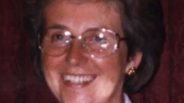 A middle-aged woman looks at the camera, she has short greying hair and glasses.