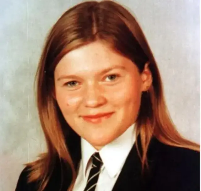 A young girl in a school uniform during a school picture. She has shoulder-length blond/brown hair and green-grey eyes. She is wearing a black blazer, white shirt and yellow-striped black tie.