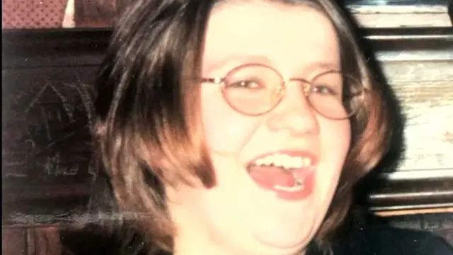 A woman with short hair and round glasses looks into the camera. She has her mouth open in a wide smile. There is wood-panelling in the background of the shot.