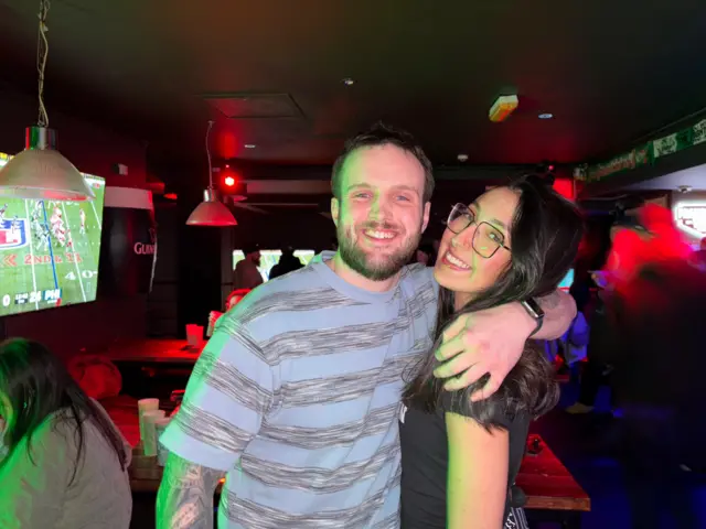 A photo of JOsh, a man in a blue and grey stripey top with an arm tattoo smiling at the camera with a beard. He's hugging Monica who has long black hair - wearing glasses and a black top.