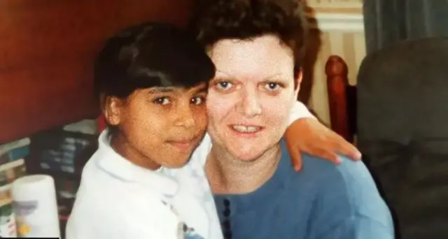 A woman and a young girl look at the camera - they are inside a house in what appears to be a living room. The woman has short hair and a blue top, the young girl is wearing a white top and has dark hair and dark skin.