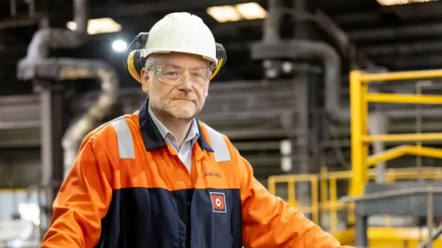 James Brand wearing protective clothing including a high vis jacket, hard hat and ear defenders with warehouse behind him