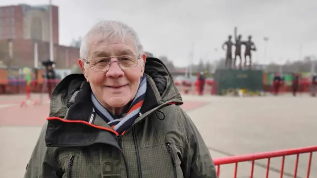 Peter Byrne wearing a coat outside Old Trafford with the statue of the 'Holy Trinity' of club legends seen in the distance