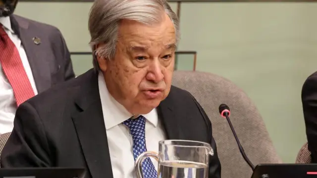 Antonio Guterres in suit and blue tie sat behind desk with microphone and water