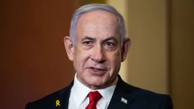 Headshot of Netanyahu showing from his shoulders up, wearing a black suit jacket, white shirt and a red tie. He has grey hair and large ears. He has an Israeli flag pin on his jacket