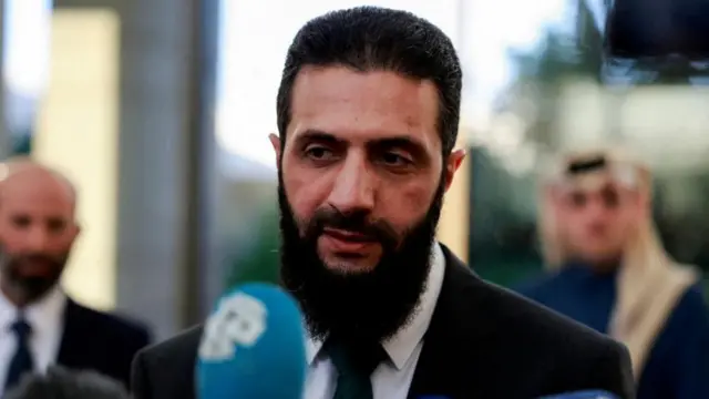 Ahmed al-Sharaa, in a suit, with reporters' microphones in front of him