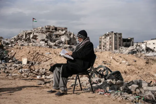 A man sits on a chair, reading a book, surrounded by rubble