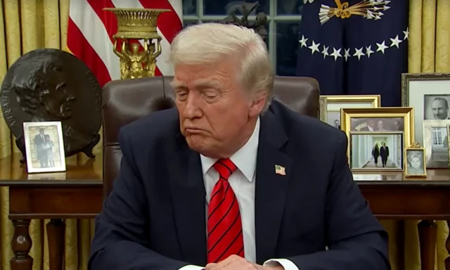 Trump sitting at a desk resting his arms on it. There's a table behind him with various family pictures and ornaments. There's also an American flag which is partly visible. Trump wears a navy suit, a badge of the American flag, a black and red striped tie and has white fluffy hair.