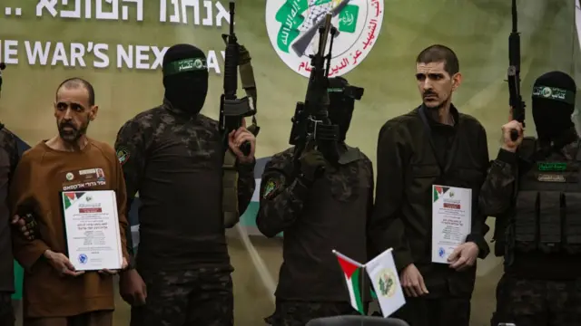 Two men in plain clothes stood in between masked man in camouflage outfits holding guns