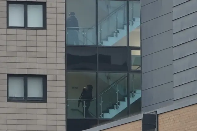 An armed police officer stands on a stairwell of an apartment block, pictured through the window