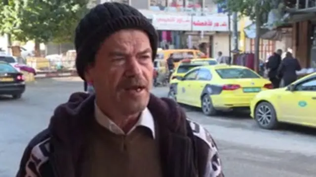 Man speaking into camera wearing hat, scarf and jumper with a road, cars and people behind