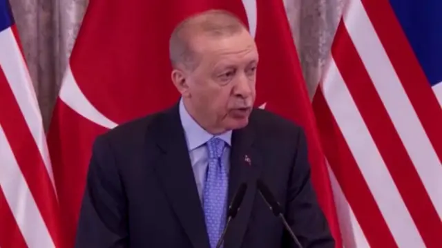 Erdogan speaking into two small microphones in front of a Turkish flag. He wears a suit jacket, a blue shirt and a blue tie