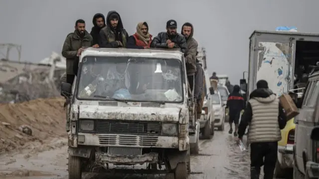 A group of people in a truck as a line of cars is behind and beside them on a busy road, some people walking on foot