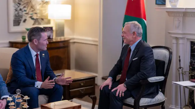 National security advisor Mike Waltz sitting down on cream-coloured couch in dark grey suit and burgundy tie as he speaks to King Abdullah II of Jorda, also sitting in a cushioned cream chair. The Jordanian flag is behind the kind, and there's a white stone fireplace visible behind him