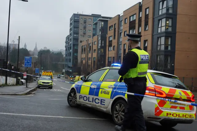 A police officer, pictured from behind, stands between a police cordon and a police car. He stares into the middle distance.