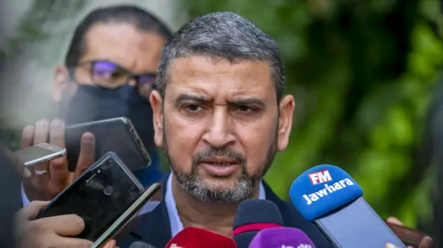 A man with a grey and black beard, moustache and hair, being surrounded by press microphones and mobile phones which are held up to his face.