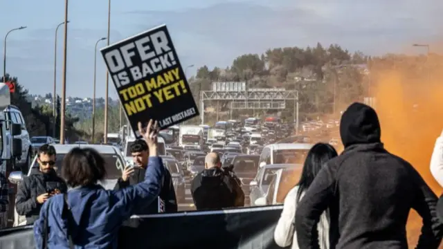 Protesters block a multi-lane highway. They hold up signs and let off orange smoke while cars can be seen queue in the distance