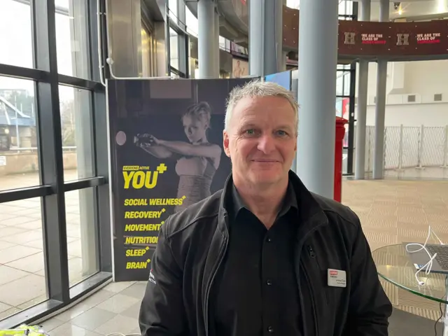 Lorenzo Clark, a middle-aged man wearing a black jacket, black shirt and a name badge. He is standing inside the foyer of a large public building.