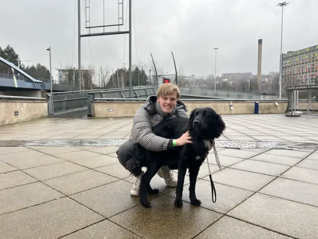 A fair-haired man in his mid 20s crouches to pet a placid-looking black dog