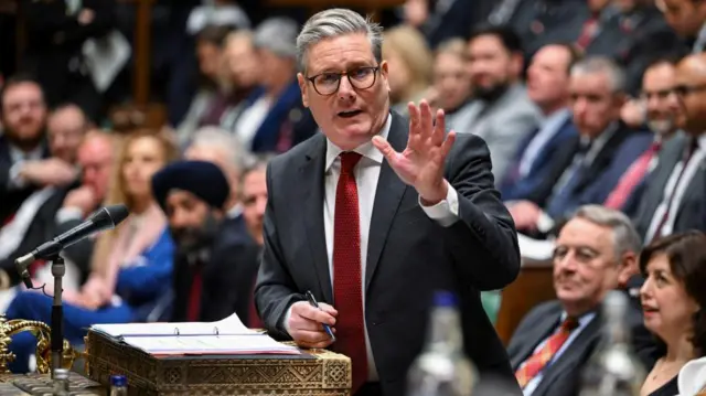 Keir Starmer stands at the dispatch box during Prime Minister's questions. He gestures with one hand, and holds a pen in the other, as he addresses MPs