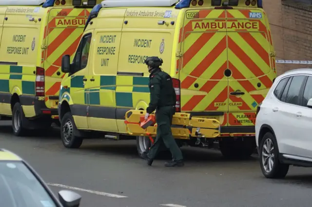 A paramedic in green overalls carries a stretcher alongside a parked ambulance vehicle.