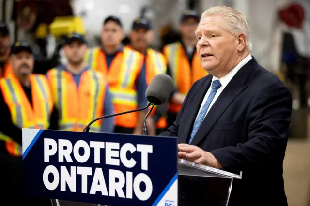 Ford stands in front of a podium with workers in orange vests behind him