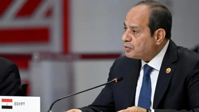 Egypt's President Abdel Fattah al-Sisi in black suit with blue tie sat at desk with microphone, and small placard with Egypt written on it.