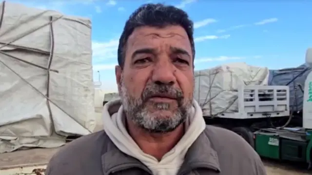 Headshot of Hussein, who has stubbly grey and black facial hair, looking into the camera. He wears a cream hoodie and grey jacket. You can see a few trucks piled high and covered in tarp.