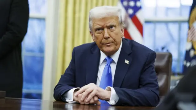 US President Donald Trump sits behind the Resolute Desk in the Oval Office. He is wearing a blue suit and US flag pin.