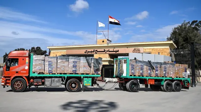 A red flatbed lorry is loaded with cases of humanitarian aid. There are stacks of cardboard boxes wrapped in plastic on the back of the lorry. Behind it you can see the Rafah border entrance, which has its name written in Arabic. The Egyptian flag flies above it. It's a sunny day with blue skies above.