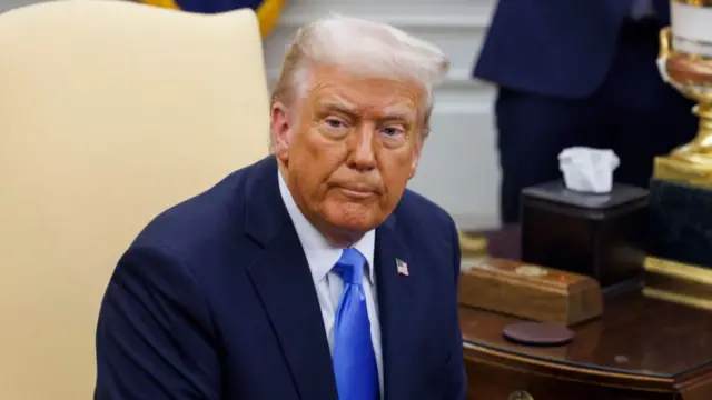 US President Donald Trump sitting on a chair in a suit with wooden desk behind him