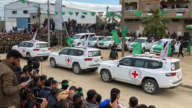 Three red cross vehicles are shown in Deir Al-Balah ahead of a hostage release last weekend. There are crowds of onlookers waiting to see the release. Green Hamas flags can be seen lining the roadway.