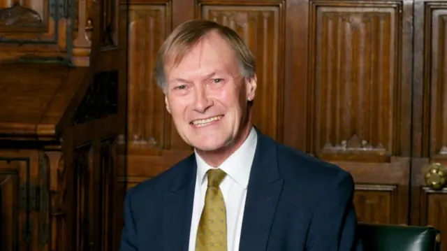 Sir David Amess smiling in front of a wood panelled wall