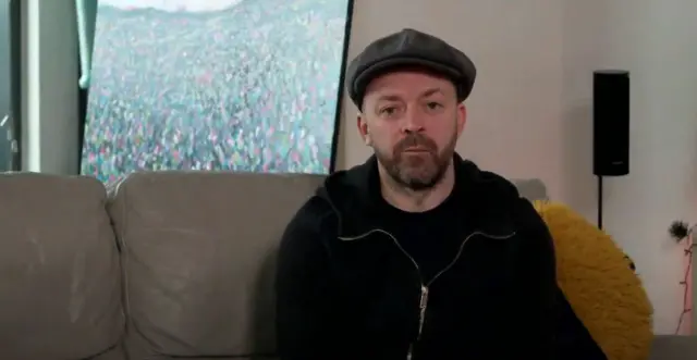 A man, wearing a grey hat and a black hoodie, sitting on a cream coloured sofa.