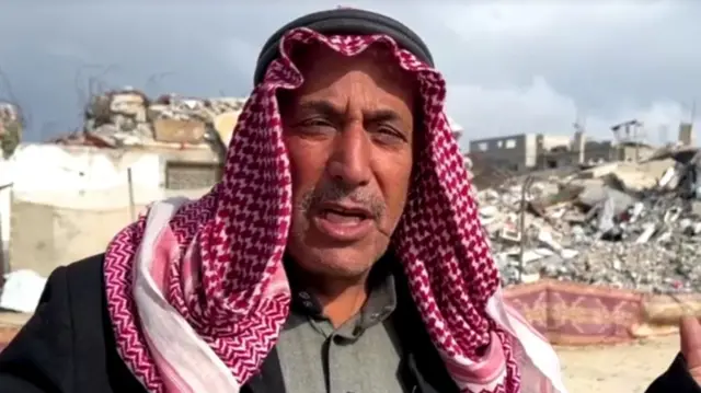 A headshot of Taha mid speech. He wears a red and white head covering secured by a black band around his head, and there's rubble behind him