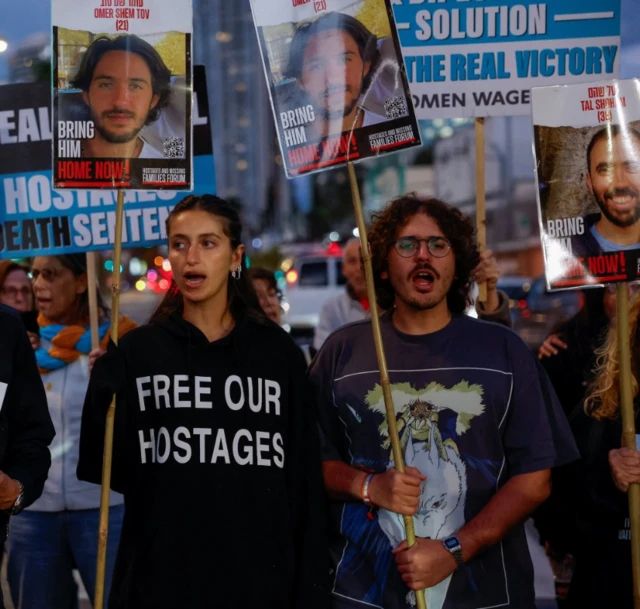 Dana Shem Tov holding a sign with the face of Omer Shem Tov, wearing a hoodie which says 'FREE OUR HOSTAGES'