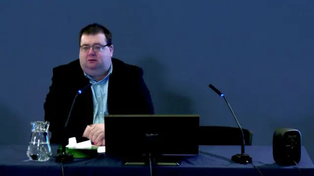 A man with black hair, wearing glasses, a black jacket and blue shirt. He is sitting at a desk with a computer in front of him. The wall behind him is blue.