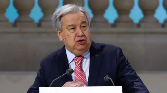 Antonio Guterres in a blue suit and red tie behind a white podium with microphones.