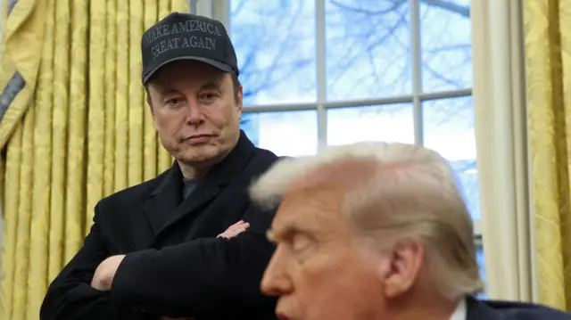 Elon Musk stands with his arms folded wearing a cap reading 'Make America Great Again'. He looks towards Trump who is seating in the foreground. The president's face is slightly blurred as he faces to the side of the camera